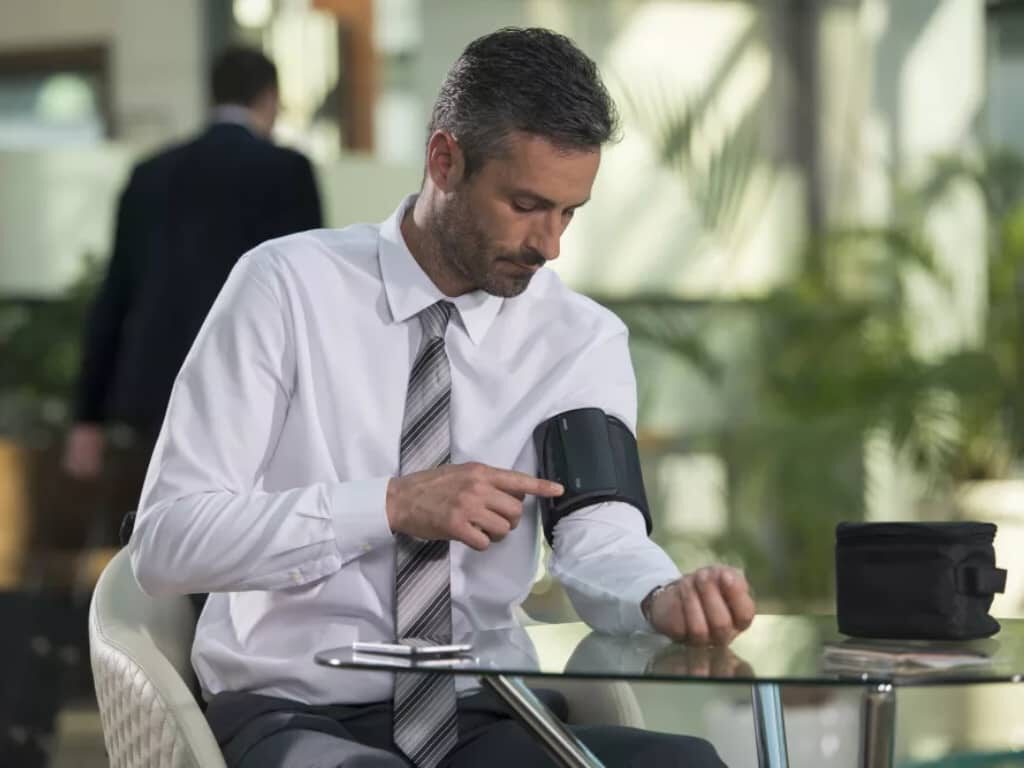 Man zit aan een glazen tafel en start een bloeddrukmeting met de Omron Evolv om zijn bovenarm, met een opbergetui en smartphone op tafel