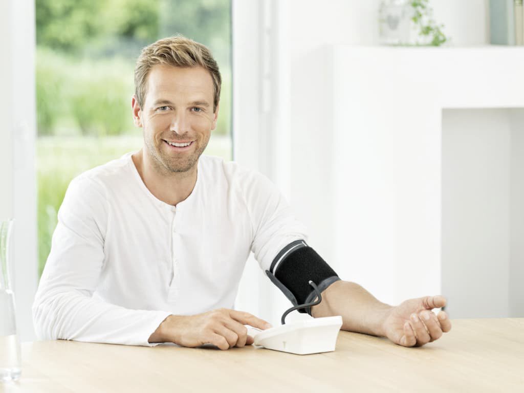 Man meet zijn bloeddruk aan tafel met de Beurer BM 26 bovenarm bloeddrukmeter en manchet om de arm