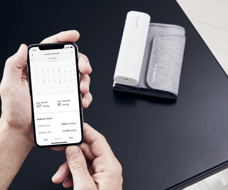 Persoon met smartphone waarop de Withings-app zichtbaar is, met de Withings BPM Connect intelligente bloeddrukmeter met wifi op tafel