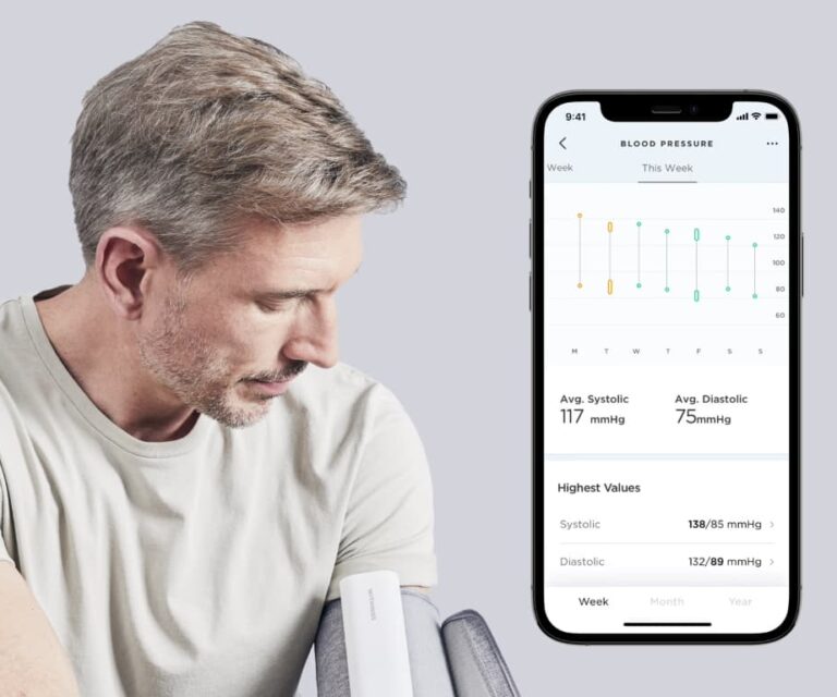 Man meet zijn bloeddruk met de Withings BPM Connect intelligente bloeddrukmeter met wifi, met smartphone groot in beeld waarop de Withings-app zichtbaar is