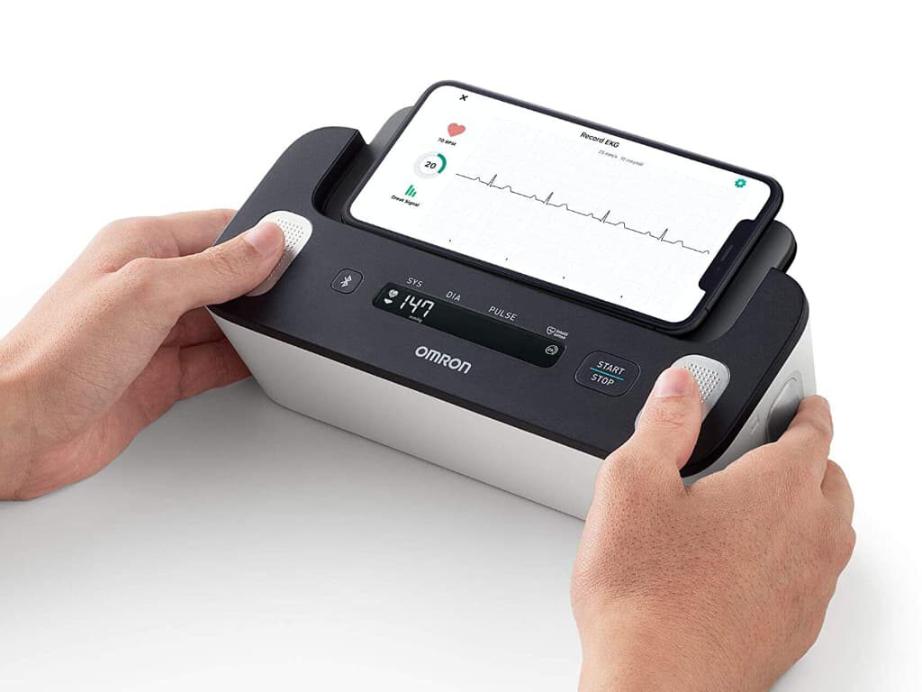 Persoon plaatst beide duimen op de Omron Complete om een ECG te meten, met een smartphone op het apparaat waarop het elektrocardiogram zichtbaar is