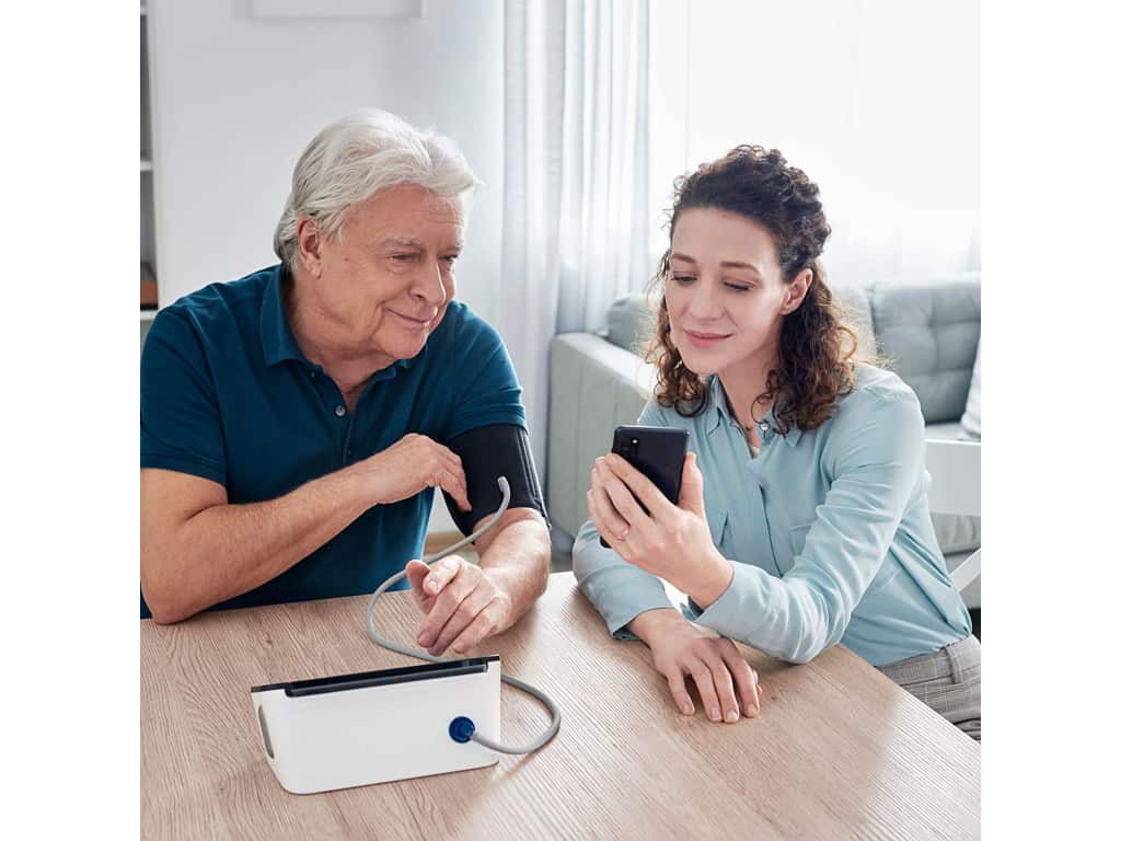Man meet zijn bloeddruk met de Omron Complete zichtbaar vanaf de achterkant, terwijl een vrouw een smartphone vasthoudt