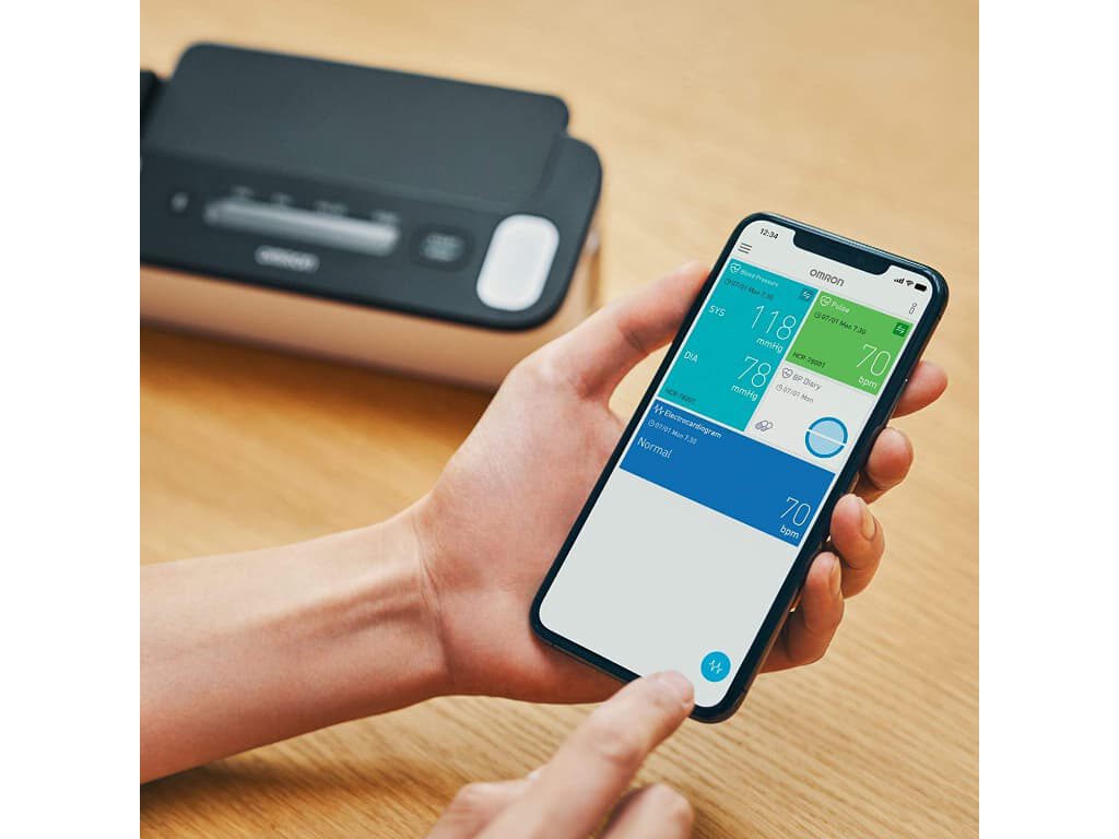 Smartphone met de Omron Connect-app in de hand op de voorgrond, met vervaagd op de achtergrond de Omron Complete bovenarm bloeddrukmeter met ECG