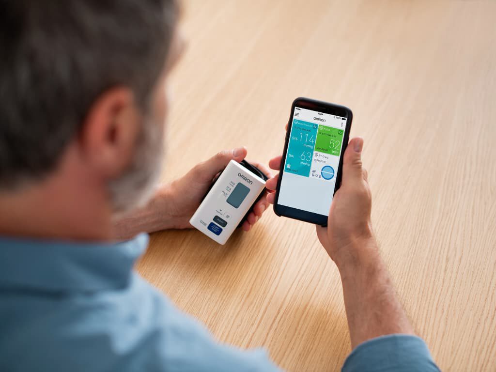 Man houdt de Omron Nightview polsbloeddrukmeter in zijn linkerhand en een smartphone waarop de Omron Connect-app zichtbaar is in zijn rechterhand