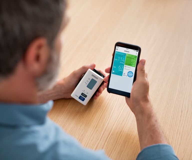 Man houdt de Omron Nightview polsbloeddrukmeter in zijn linkerhand en een smartphone waarop de Omron Connect-app zichtbaar is in zijn rechterhand
