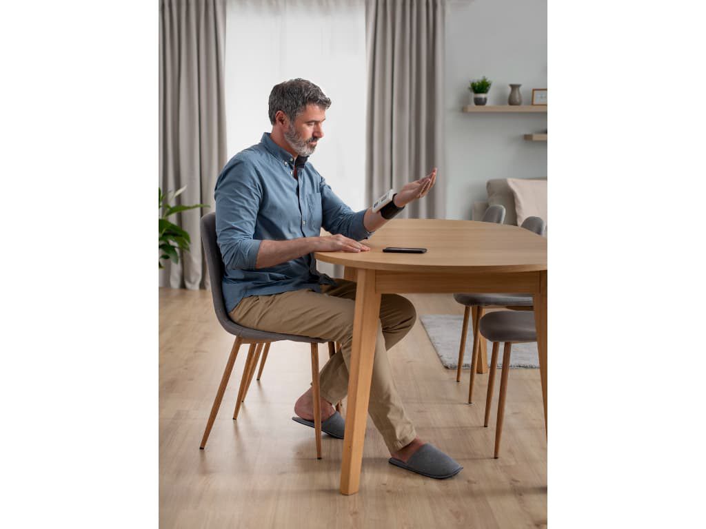 Man zit aan tafel en meet zijn bloeddruk met de Omron Nightview polsbloeddrukmeter, met een smartphone op tafel