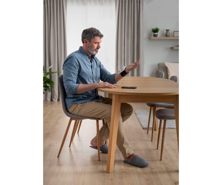 Man zit aan tafel en meet zijn bloeddruk met de Omron Nightview polsbloeddrukmeter, met een smartphone op tafel