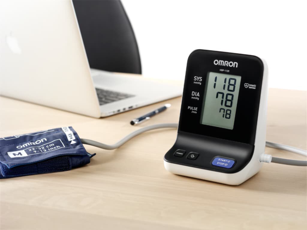 Omron HBP-1120 professionele bloeddrukmeter met aangesloten manchet maat M op tafel, met op de achtergrond een laptop en een pen