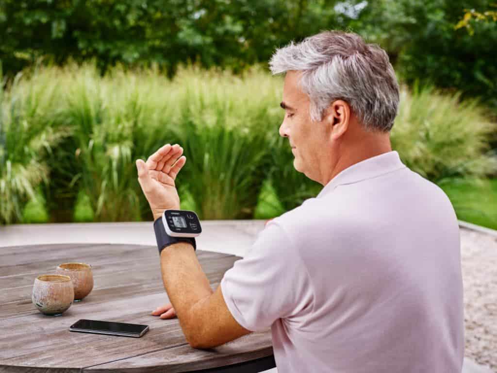 Man zit aan tafel met de Omron RS3 Intelli IT polsbloeddrukmeter om zijn pols en een smartphone op tafel