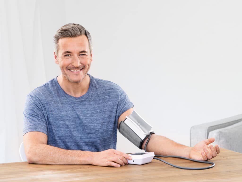 Man meet zijn bloeddruk aan tafel met de Beurer BM 51 easyClip bovenarm bloeddrukmeter en easyClip-manchet om de arm