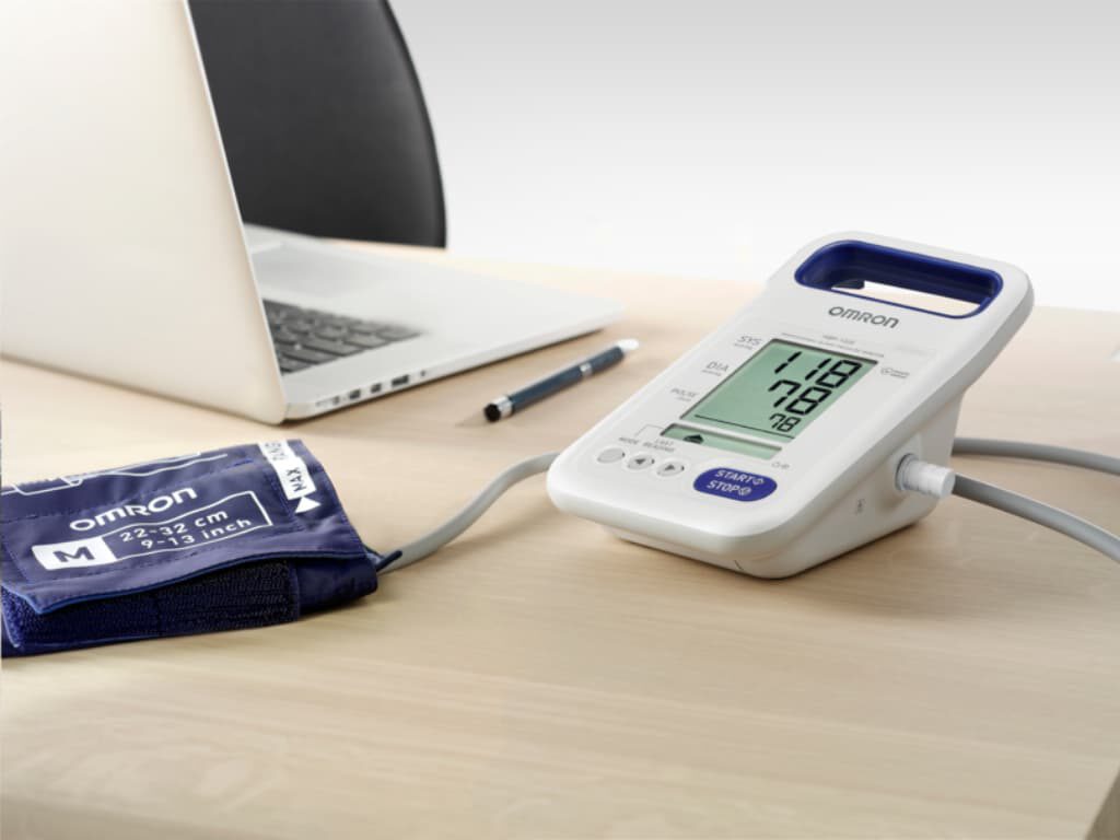 Omron HBP-1320 professionele bloeddrukmeter met aangesloten manchet maat M op tafel, met op de achtergrond een laptop en een pen