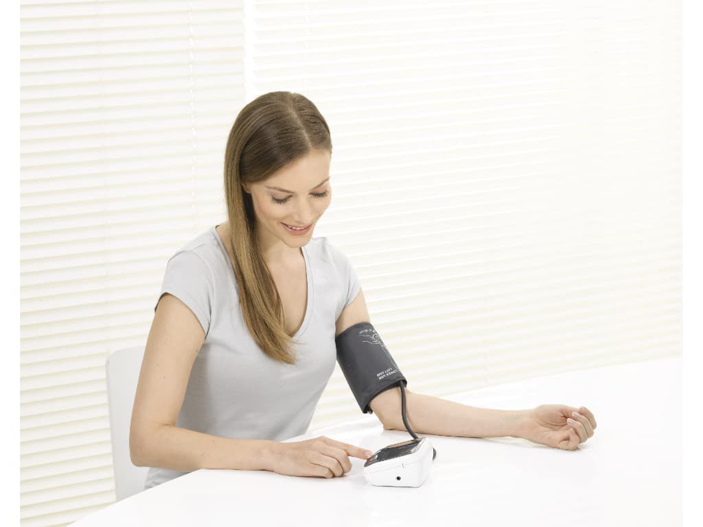 Vrouw meet haar bloeddruk aan tafel met de Beurer BM 40 bovenarm bloeddrukmeter en manchet om de arm