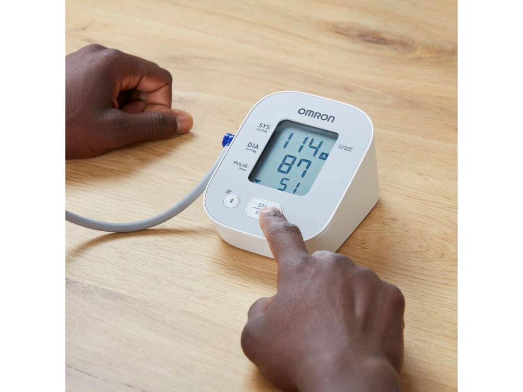 Omron X2 Smart+ bovenarm bloeddrukmeter op tafel met meetgegevens op het scherm en vinger op de start/stop-knop